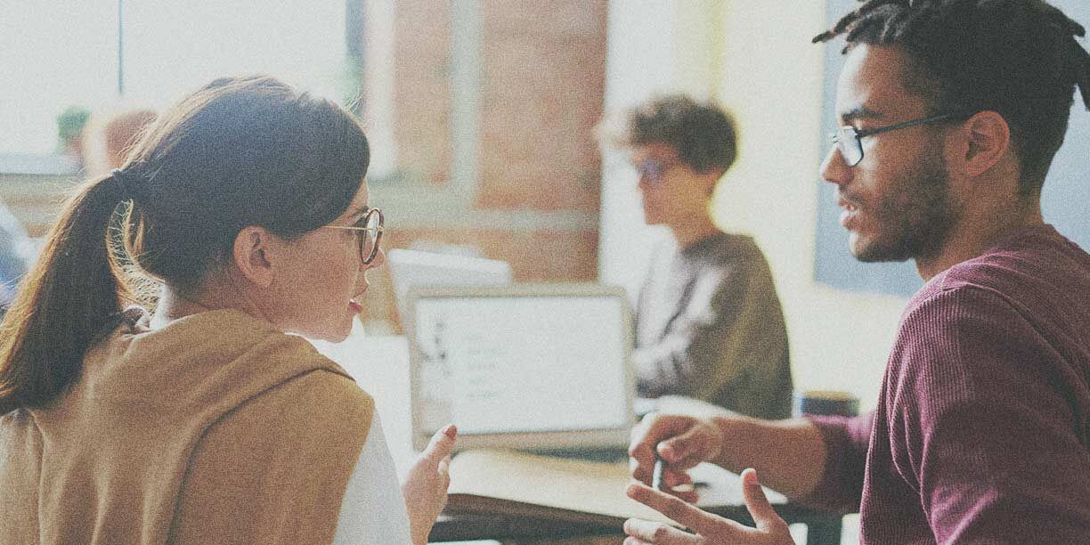 woman with brown hair and tan sweater talking to a man with glasses in an office