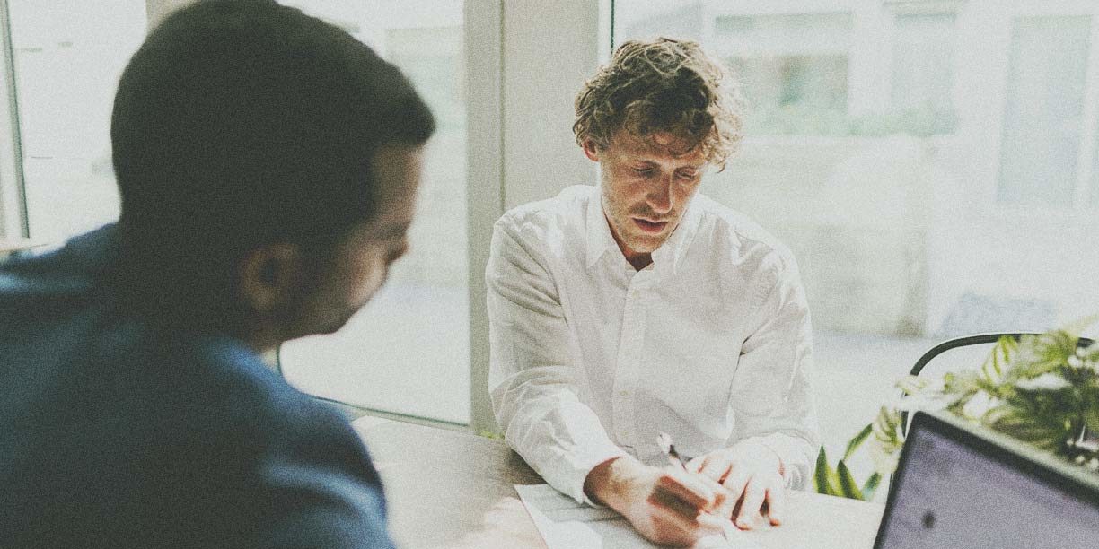 Man in white writing on a piece of paper with a man in dark blue watching