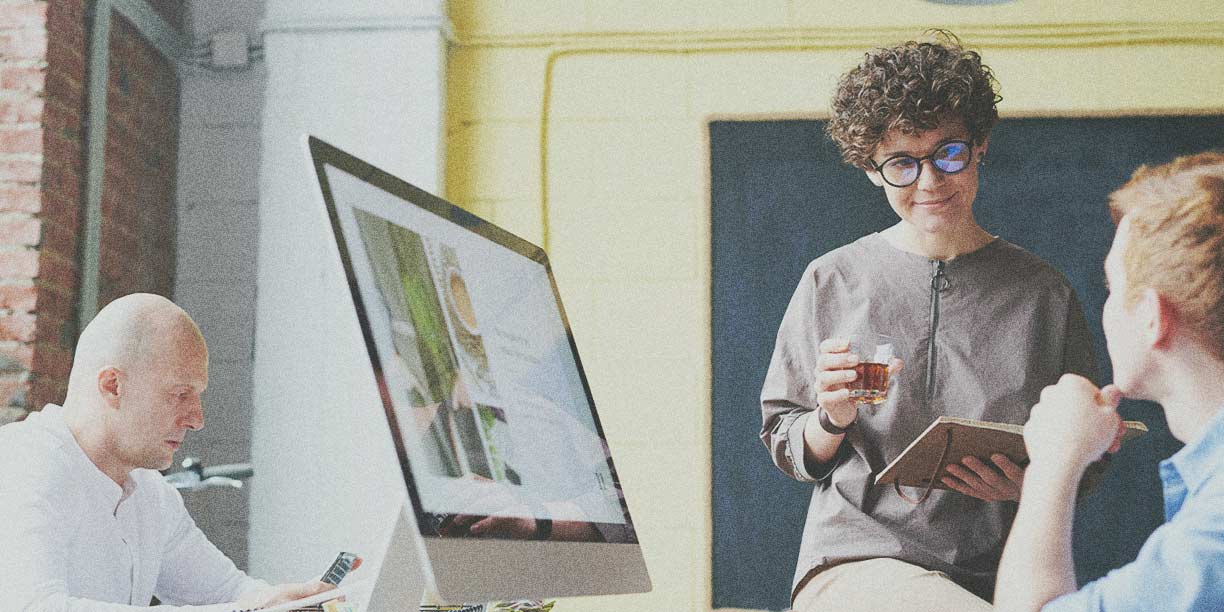 Woman in light brown holding a cup of coffee working at a computer with man in blue